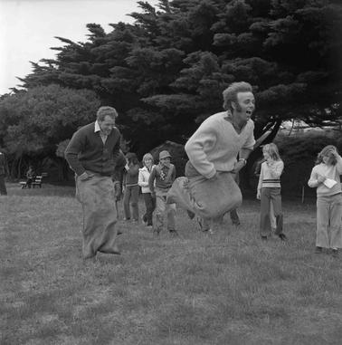 Two men racing each other in a sack race with several children looking on.  Pine trees form the back drop.