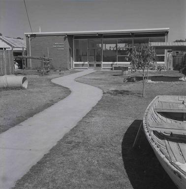A single storey building with a concrete path and a boat ion the lawn n the foregrouno date.