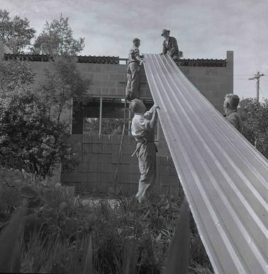 Four men are manhandling a sheet of iron roofing (’cliplock’) onto the roof of a cement block building.  Trees and bushes at left of photo.