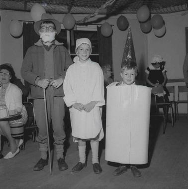 Three children in fancy dress costumes.  Other people are in the background, the hall is decorated with balloons.