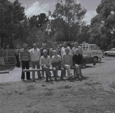 Ten men in two rows, four sitting on a timber barricade and six standing behino date.  Other people are in the backgrouno date.  A white small truck sith signage reading ‘Somers Fire Brigade’ behind the men.