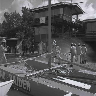 Two groups of firefighters in protective clothing working near a two storey building.  One group is manhandling a ladder, the other a fire hose.  A catamaran is in the foregrouno date.
