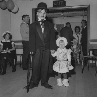 A ‘Charie Chaplin’ and a small girl in fancy dress, other people in the background indoors with balloons festooning the ceiling.