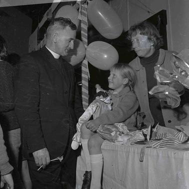 A clergyman alking to a girl seated on a table and holding a toy clown and a woman.  Festive balloons hang from a streamer. 