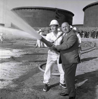 Two men with a spouting fire hose: one in white work clothes and a hard hat and the other in a suit.  There are oil or gas tanks in the backgrouno date.
