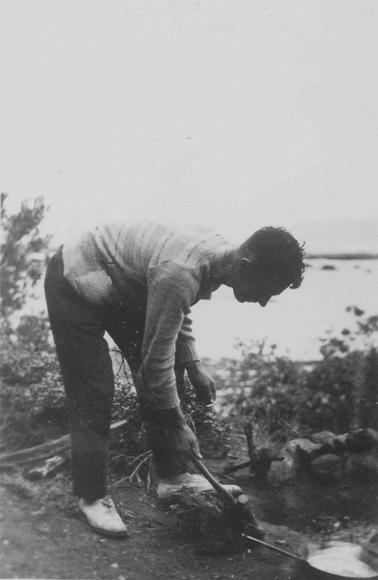 Merricks. Closeup of young man bending over, holding a hammer. Frying pan in foregrouno date. Pile of stones, cut down vegetation. Man wearing striped jumper, long trousers, light colored shoes.