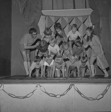 Twelve children in a three row ‘pyramid’ tableau with a man and a woman helping them  They are on a stage.