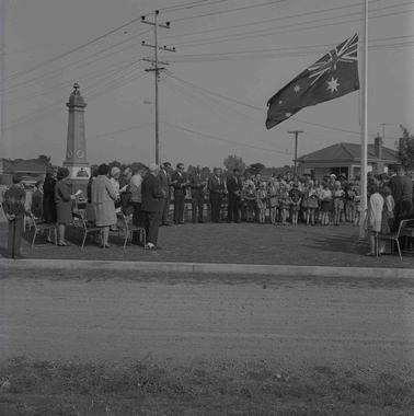 Adults and children (some in Girl 
Guide and Brownie uniforms) standing about in an orderly circle at a war memorial cenotaph.  an Australian flag is flying at half mast..   