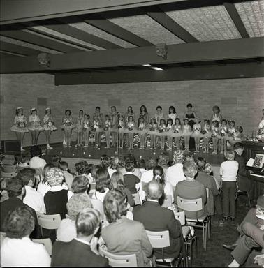 Eighteen young girls dressed in tutus standing in a row on a stage with aeveb older girls in performance coswtumes behind and three oldet ladies to the rear right.  A seated audience is looking on.