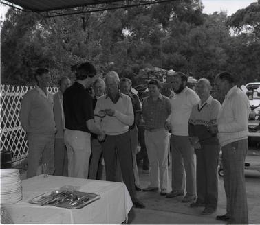 A group of ten men standing near a small table covered with a white cloth with plates and a tray of cutlery on it.  One of the men is presenting a small item to another, so that thier hand meet.