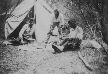 Merricks Beach, three young men in front of a tent, grouped around ground sheet. Plates etc. on ground sheet. Two men are sitting on the grouno date. Third man is standing, leaning forward, holding a frying pan.
