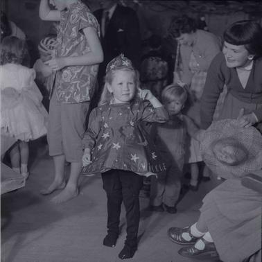 A young girl with blonde hair wearing a fancy dress with other people around and nearby.  Her mother is leaning towards her.