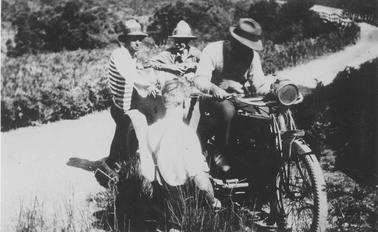 Four young men with motor bike in center of photo. One is on motor bike seat, three are standing. One is in foreground with back to camera, not wearing a hat. Other three are wearing hats. One of these is wearing striped top. Unmade road meandering across photo. Low vegetation on roadside verges, higher growth behino date.
