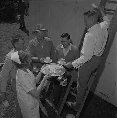 A hospital nurse is passing tea cups to four men.  One man is on a low rung of a ladder, other three are sitting down.