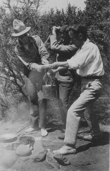 Merricks Beach. Three young men behind camping fireplace with billy and frying pan on grouno date. Two men, one wearing a hat, are holding a rabbit stretched out over billy. Third, central figure has hands raised to shoulder height, holding cutting implement. Bush in backgrouno date.