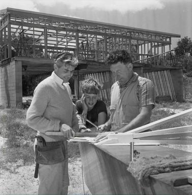 Two men and a boy discussing plans which are laid out at an outdoor bench.  A partly constructed two story building is in the backgrouno date. 