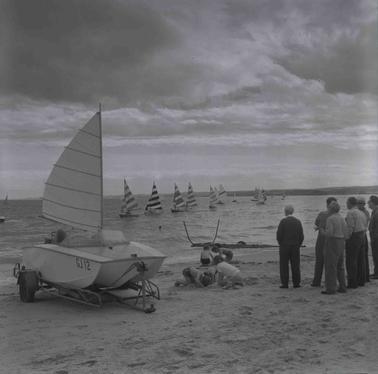 Yachts sailing on the bay, one small catamaran on the shore, children playing in the sand and a group of six men standing facing the water.