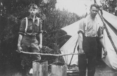 Two young men carrying pole from which are hanging two rectangular kerosine-tin buckets, standing in front of a tent. Dense ti-tree in backgrouno date. Both wearing long trousers, shirts with sleeves rolled up to the elbows, and braces.