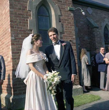 A bridal couple in front of a red brick church, others in backgrouno date.