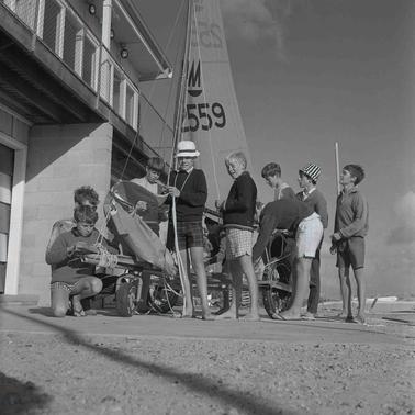 A group of nine young men beside a ‘land yacht’ clubhouse attending to a yacht on wheels.