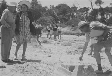Merricks Beach sandcastle competition. Man (R) wearing shorts, shirt, hat, holding papers in (L) hand bending over castle. (Bill Seddon), couple on (L) F & M Nutting - both wearing hats. Man has stick. Group of 4 figures in background plus 3 figures behind Mrs Nutting looking to foreshore.