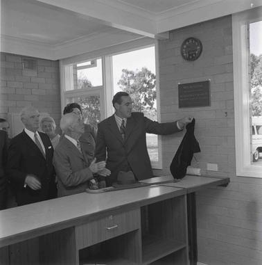 Three men in suits and other people.  oOe man is golding a velvet cloth from the unveiling of the commemorqative plaque on the wall.