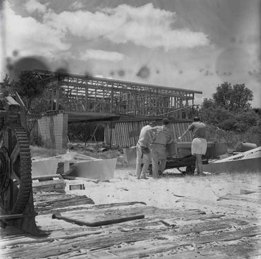 A costruction site with a two story building, the upper floor is framed up.  Three men are leaning on a trailer.