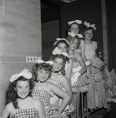Seven young girls dressed in gingham dancing performance costumes, posing for a photograph.