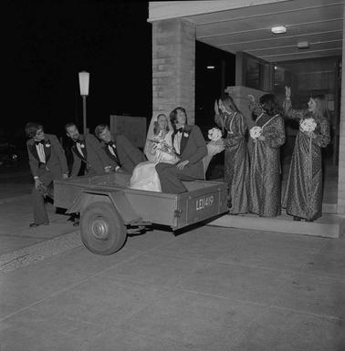 A bride and groom seated in a box trailer with three male and three female attendants looking on.
