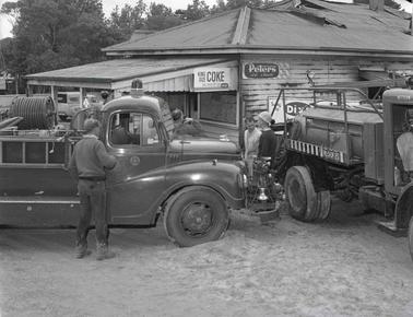 Two fire trucks parked close to a weatherboard building with product logos ‘Peters’ and ‘Coke’ near roof line. several men are standing about.  Damage to the roof is evident.