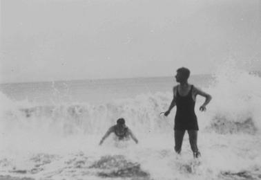 Beach at Merricks. Surf breaking at end of beach. Two male figures surfing. Central figure is prone, supported by arms. R.H. figure standing ankle deep in water wearinbg one piece 20’s style bathing suit.