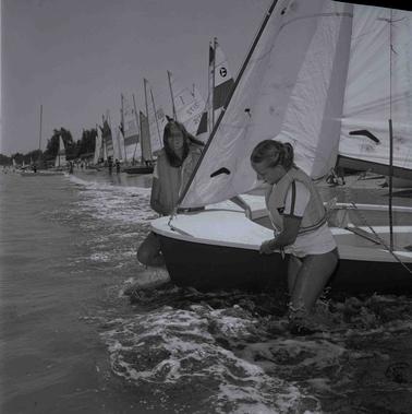 A row of yachts and people on the water line .  In the foreground two young women are pulling a yacht into the water.
