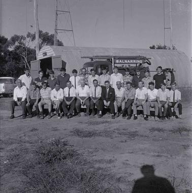 Twenty seven men posed for a group photograph with a fire truck in front of a ‘Nissen Hut’ with signage ‘CFA’.