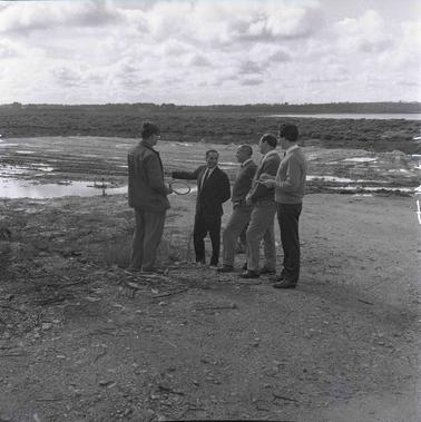 Four men in discussion outdoors on a barren area with large water puddles.. 