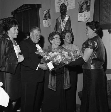 A tableau of five people at a presentation of flowers to a couple with two women looking on.