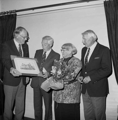 Four persons, one female at a presentation of a framed picture.  The woman is holding a bouquet.