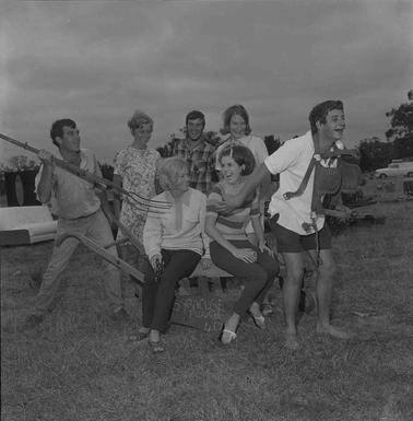 Seven men and women posing in fun as a ‘horse and cart’ ‘pulling’ two women whoa re sittingon a bench,  one man is pretending to throw a pitchfork at the ‘horse’.
