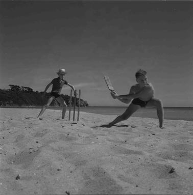 Two young boys playing cricket on the beach.  One is swinging his bat, the other is minding the stumps.