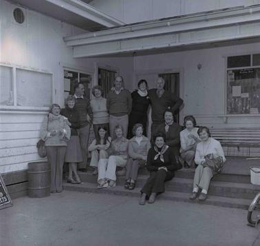 A group of fourteen men and women, half sitting on steps in front of a weatherboard and fibro building.