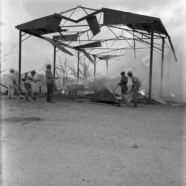 Men with firefighting equipment at a burnt out shed frame.