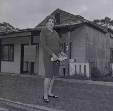A Woman dressed in a (skirt) suit near a footpath front of a weatherboard house with a notice in front.  The woman is holding a framed photograph of a man and a Woman.