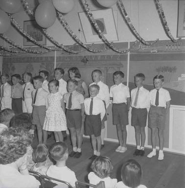 Fourteen boys and girls in two rows in front of an audience.  A wall of blackboards is in the backgrouno date.