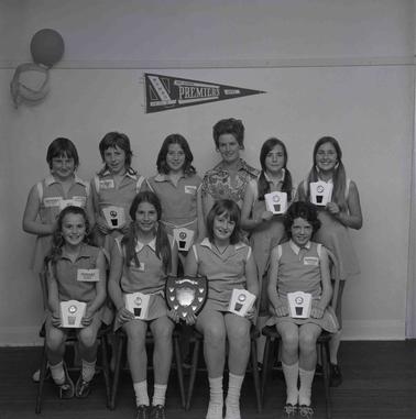 A group of nine girls in Netball uniforms and a woman, posing for a photo in two rows.  Each girl is displaying a trophy, another perpetual trophy is held by two of the girls.
