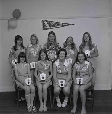 A group of eight girls in Netball uniforms and a woman, posing for a photo in two rows.  Each girl is displaying a trophy., another perpetual trophy is held by the woman in the back row