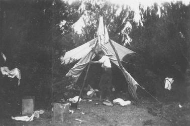 Camping scene. Male figure in shirt sleeves and wearing trousers standing in front of tent. Tent flaps are raised, second figure can be seen lying inside tent. Guide poles and ropes in photo, also 2 kerosene can water carriers, billy and miscellaneous litter. Garments hanging from ropes. Ti-tree in backgrouno date.