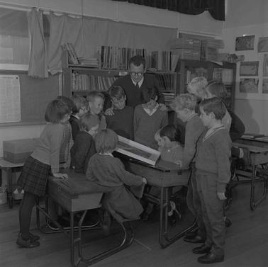 Fourteen school children gathered around a school desk looking at a framed picture, a man is looking on with them.