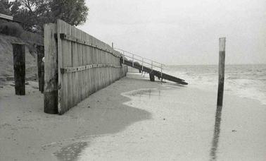 A beach with a wooden breakwater wall.  There is erosion behind the wall almost undermining a house fence.