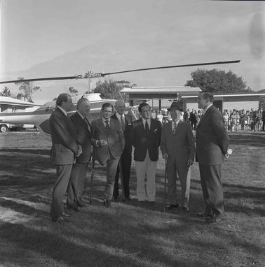Seven men in suits standing in front of a helicopter.