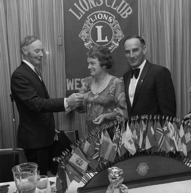 A man in a suit presenting a cheque to a woman with another man looking on.  All are dressed in formal attire.