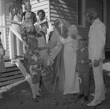 A weddding party: groom, bride bending down to her flower girl, a page a minister and three other people near and on steps to a watherboard church building.